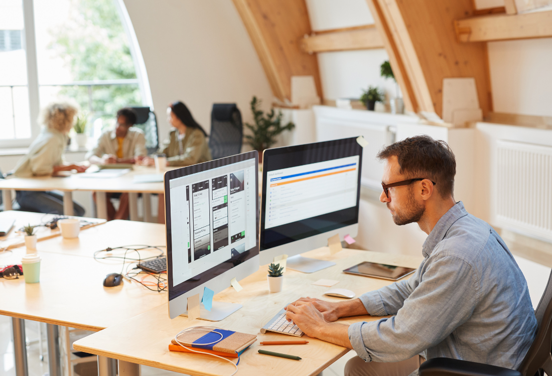 Man working at dual monitors in a modern office while colleagues discuss in the background, representing SaaS solutions for finance teams.